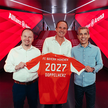 Trois hommes souriants se tiennent sous l’enseigne de l’Allianz Arena, tenant une maillot de football rouge du FC Bayern Munich. Le maillot affiche "2027" et "DOPPELHERZ" au dos. L'atmosphère est festive, suggérant un événement important pour le club.