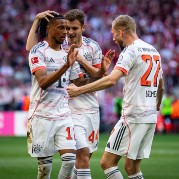 Quatre joueurs de football en maillots de Bayern Munich célèbrent un but. L'un d'eux, avec des dreadlocks, lève les mains en signe de joie. Les autres sourient et s'approchent pour le féliciter. L'ambiance est visiblement festive au stade.