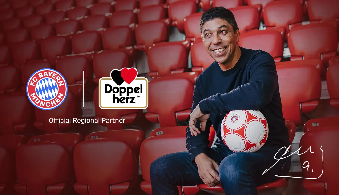 Un homme souriant assis dans des sièges rouges, tenant un ballon de football. Logo du FC Bayern Munich et celui de Doppelherz présents sur l'image, indiquant un partenariat. L'ambiance est conviviale et en lien avec le sport.