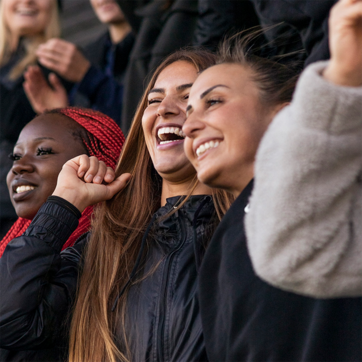 Trois femmes souriantes affichent leur joie, levant les mains en signe d'encouragement. Elles portent des manteaux noirs et un haut gris, et sont entourées d'autres personnes en arrière-plan. L'image transmet une ambiance festive et d'excitation.