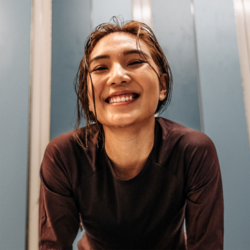 Une jeune femme souriante, les cheveux mouillés, porte un maillot de sport noir. Elle est penchée en avant, exprimant une attitude joyeuse et dynamique, dans un cadre avec des murs aux teintes claires.
