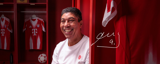 Un homme souriant est assis dans un vestiaire aux murs rouges, avec des maillots de football affichés derrière lui. Une balle de football est visible sur le sol. Son nom est écrit en bas à droite.
